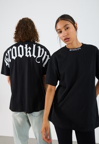 Svart oversized t-shirt med "Brooklyn" i fet vit gotisk stil på baksidan och en mindre version på framsidan vid kragen. Tillverkad av bomullsmaterial.