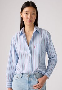 Chemise boutonnée à rayures bleues avec des lignes verticales rouges, à manches longues et une poche poitrine. Tissu doux avec une coupe décontractée. Associée à un denim clair.