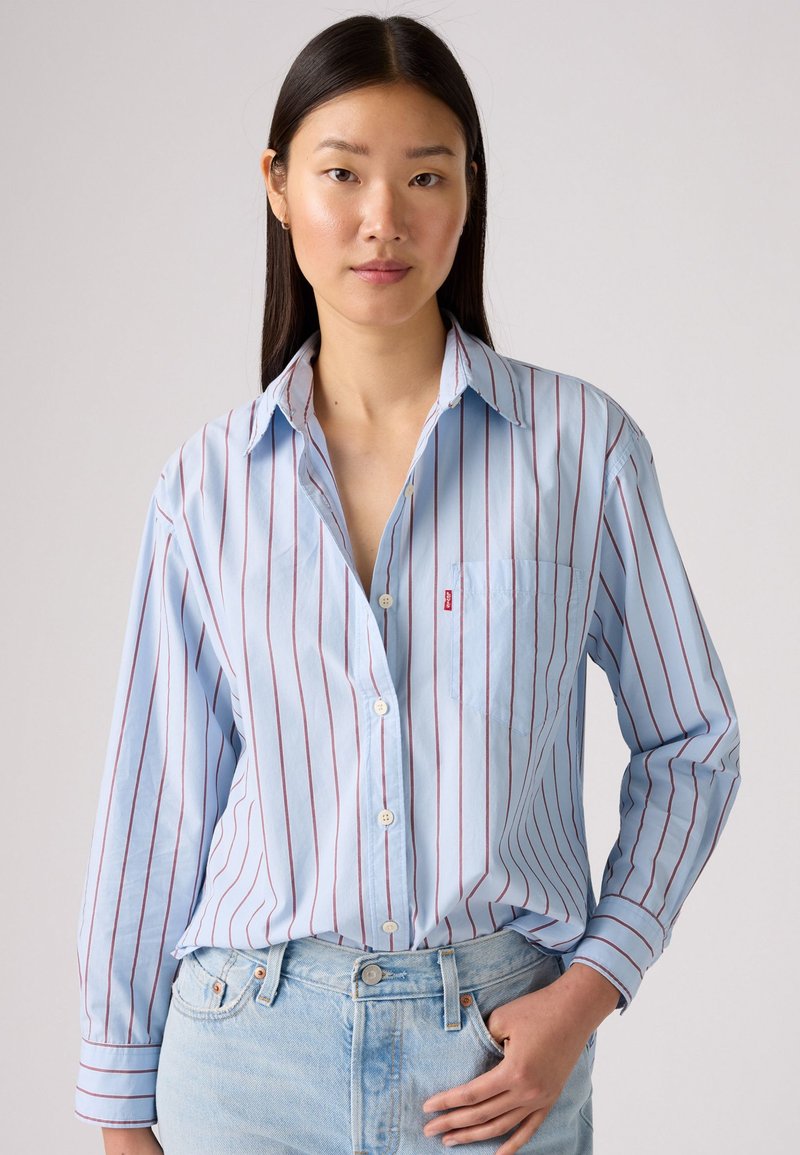 Chemise boutonnée à rayures bleues avec des lignes verticales rouges, à manches longues et une poche poitrine. Tissu doux avec une coupe décontractée. Associée à un denim clair.