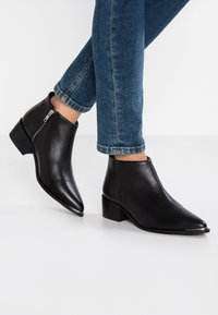 Schwarze Knöchelstiefel mit spitzen Zehen und niedrigen Blockabsätzen. Aus strukturiertem Leder gefertigt, mit einem Reißverschluss an der Außenseite.