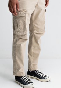 Man in beige cargobroek en zwarte hoge canvas sneakers staat op een lichte vloer met een effen achtergrond.