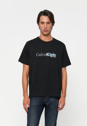 Jonge man in een zwart Calvin Klein New York logo t-shirt en blauwe jeans, staand tegen een effen witte achtergrond.