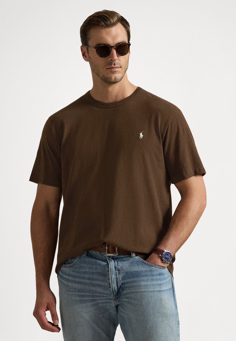 Καφέ βαμβακερό μπλουζάκι με κοντά μανίκια και στρογγυλή λαιμόκοψη, με μικρό λευκό λογότυπο στο αριστερό στήθος, συνδυασμένο με ανοιχτό μπλε τζιν.