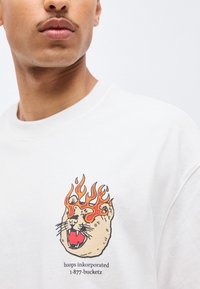 Άνδρας που φοράει λευκό πουκάμισο με γραφικό κεφάλι γρυπαλού γάτου περιτριγυρισμένο από πορτοκαλί φλόγες και κείμενο «hoops inkorporated 1-877-bucketz» από κάτω.