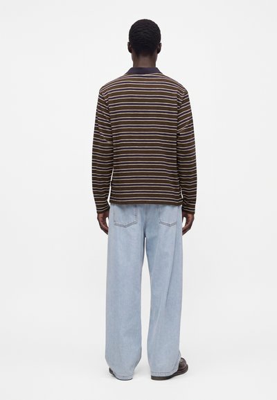 Homme debout de dos, portant une chemise à manches longues rayée marron et blanche et un jean large bleu clair avec des chaussures foncées.