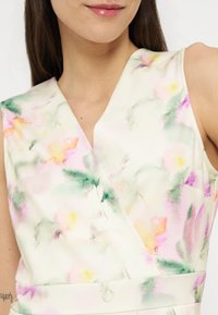 Γυναίκα που φοράει αμάνικο φόρεμα με μοτίβο λουλουδιών σε υδατογραφίες και απαλά παστέλ αποχρώσεις ροζ, πράσινου, κίτρινου και μοβ, με V-λαιμόκοψη και εφαρμοστή μέση.