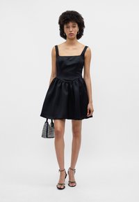 Melna satīna kleita ar kvadrātveida izgriezumu, biezām siksnām un saburzītu svārku. Valkāta ar siksnu augstiem papēžiem un mazu, mirdzošu somiņu.