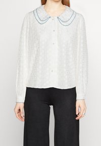 White long-sleeve blouse with a floral texture, oversized rounded collar edged in blue, and button-up front. Worn with black pants.