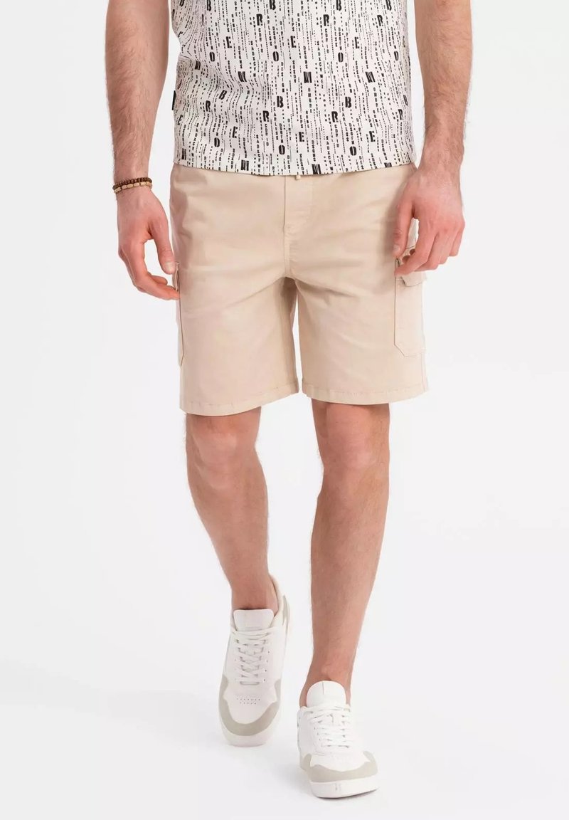 Shorts cargo beiges avec poches latérales, en coton, devant plat et coutures surpiquées, associés à des baskets blanches et une chemise à motif graphique.