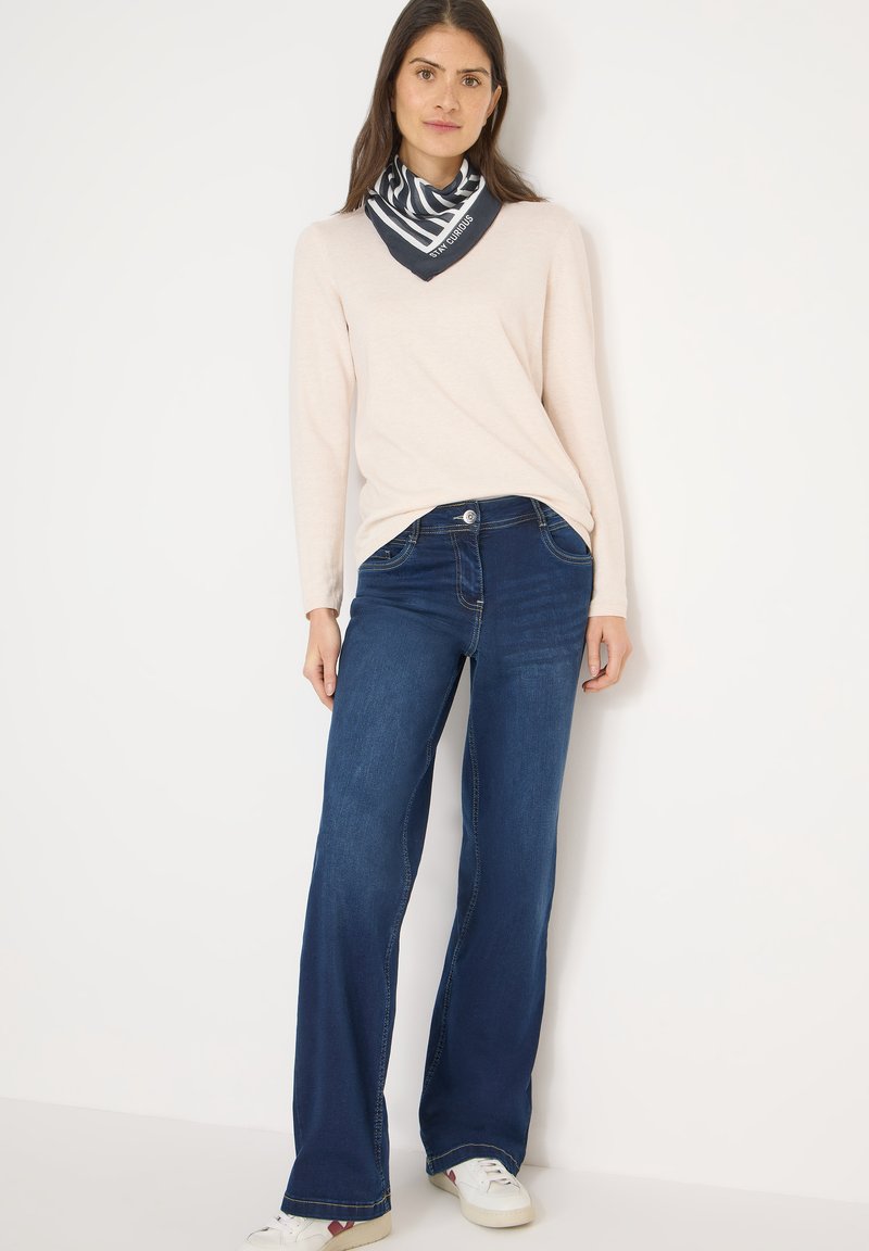 Haut à manches longues rose clair, jean évasé bleu foncé et écharpe rayée marine. La tenue présente des textures douces et des baskets décontractées.