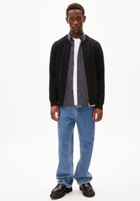 Zwarte bomberjack over een gestreepte blouse, witte T-shirt en blauwe jeans. Dikke zwarte schoenen. Eenvoudige, casual outfit met gelaagde texturen.