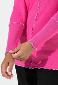 Vaaleanpunainen pitkähihainen paita, jossa on röyhelöreunat ja sininen kontrastitikkaukset, näyttäen teksturoidun kankaan. Kädet säätävät hihansuun yksityiskohtaa.