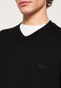 Gros plan d'une personne portant un pull noir en V par-dessus une chemise blanche, avec « BOSS » brodé sur la poitrine.
