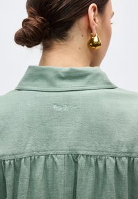 Kobieta z brązowymi włosami związanymi w kok, ubrana w jasnizieloną koszulę z małym haftowanym logo oraz dużą złotą trójkątną kolczykiem.