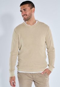 Beige gerippter Pullover mit langen Ärmeln und Rundhalsausschnitt, über einem weißen Hemd getragen. Glatte Textur mit subtiler Dehnbarkeit.