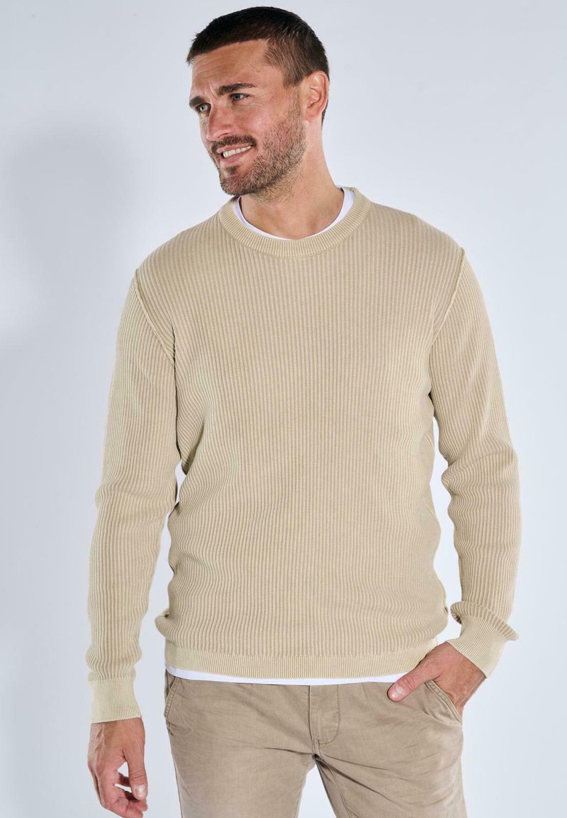 Beige gerippter Pullover mit langen Ärmeln und Rundhalsausschnitt, über einem weißen Hemd getragen. Glatte Textur mit subtiler Dehnbarkeit.