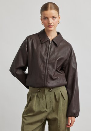 Frau mit zurückgekämmtem Haar trägt eine braune Lederjacke mit Reißverschluss und grüne, hoch taillierte Hose mit Knopfdetails vor schlichtem Hintergrund.