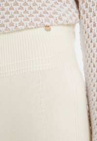 Cream-colored knit fabric with a ribbed waistband and side seam detailing, featuring a small metallic logo accent.