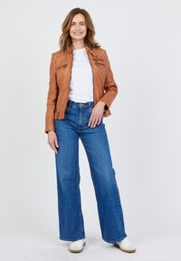 Brun läderjacka med dragkedjor, vit t-shirt, högmidjade blå vida jeans och vita sneakers. Neutral bakgrund.