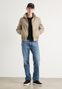 Beige bomberjakke med høy krage, kombinert med svart skjorte, blå jeans og svarte joggesko. Svart caps med hvite logo detaljer.