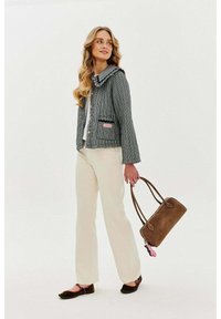 Texturovaná šedá bunda s volánkovým límcem, černými detaily a růžovou nášivkou, v kombinaci se krémovými kalhotami a hnědou kabelkou.