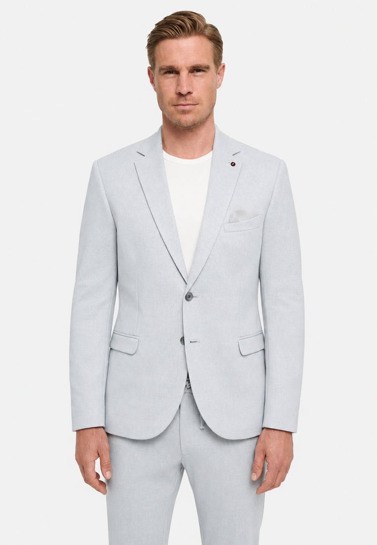 Mężczyzna ubrany w jasnoszary marynarkę i dopasowane spodnie na białym t-shircie, stojący na jednolitym białym tle.