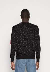 Man som ses bakifrån, iförd en svart sweatshirt med vita stjärnmönster och ljusblå jeans, mot en enkel vit bakgrund.