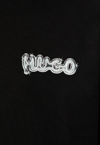 Czarna tkanina z teksturowanym logo "Hugo" w białym kolorze o błyszczącym wykończeniu i gwiazdkowymi akcentami wokół.