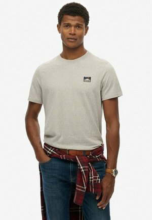 Grå bomulds t-shirt med rund halsudskæring og en lille logo-patch. Kombineret med blå jeans og en rødternet flannel skjorte knyttet om taljen.