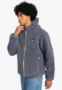 Veste en polaire gris foncé avec une finition texturée, fermeture éclair, capuche réglable et accents de zip verts contrastants. Comprend des poches latérales.