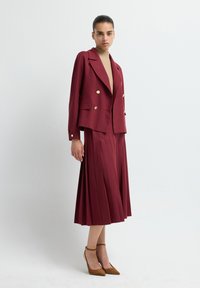 Burgundy blazer with gold buttons and pleated midi skirt, complemented by a beige turtleneck and brown heeled shoes. Smooth fabric, tailored fit.