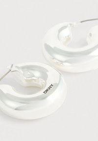 Srebrne naušnice u obliku obruča s glatkim sjajem, s uvijenim dizajnom i logotipom DKNY na jednoj naušnici. Glatka, reflektirajuća tekstura.