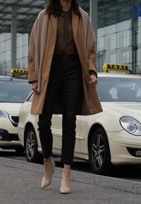 Beżowy wełniany płaszcz na leopardziej bluzce, czarne, teksturowane spodnie i beżowe botki za kostkę z zamkiem, na tle taksówki.