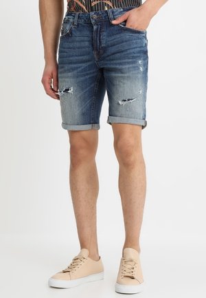 Niebieskie szorty jeansowe z podwiniętymi mankietami, z przetarciami i dopasowanym krojem. Połączone z beżowymi butami casualowymi.