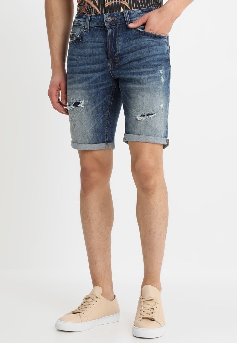 Shorts en denim bleu avec ourlets retroussés, présentant des détails usés et une coupe ajustée. Associés à des chaussures décontractées beige.