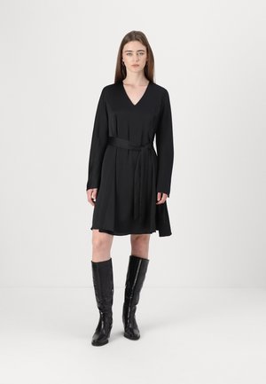 Abito nero con maniche lunghe e scollatura a V, segnato in vita da una cintura in tessuto. La modella indossa stivali neri alti e texture.