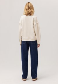 Blondynka stoi tyłem, ubrana w kremową, oversize'ową bluzę, ciemnoniebieskie spodnie dzwony i w paskowane buty typu slip-on.