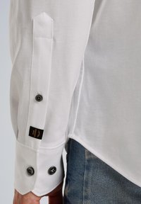 Weißes Hemd mit geknöpften Manschetten, ausgestattet mit einem schwarzen Knopf und einem Etikett. Glatte Textur und spitzes Manschettendesign. Über Jeans getragen.