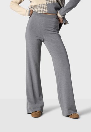 Personne portant un pantalon gris ample taille haute, des chaussures beige clair et un pull beige et gris à motifs, les mains sur les hanches.