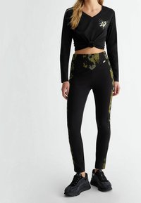 Top negro de manga larga con un nudo en el frente, combinado con leggings ajustados negros que tienen paneles laterales florales. Zapatillas negras completan el look.