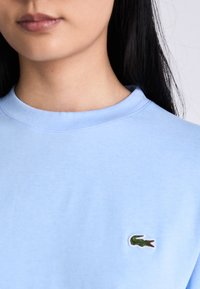 Nahaufnahme einer Person, die ein hellblaues Hemd mit einem grünen Krokodillogo auf der Brust trägt.