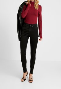 Woman wearing a burgundy long-sleeve top, black high-waisted skinny jeans, black strappy heels, holding a black leather jacket over her shoulder.