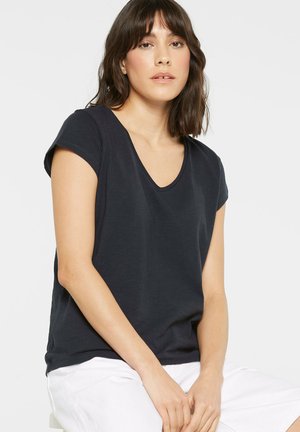 Woman with shoulder-length brown hair wearing a black short-sleeve V-neck shirt and white pants, sitting with hands resting on her lap.