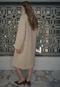 Beżowy wełniany płaszcz z wysokim kołnierzem, długimi rękawami i luźnym krojem. W zestawieniu z brązowymi szpilkami o zaostrzonych czubkach, na tle wzorzystej ściany.