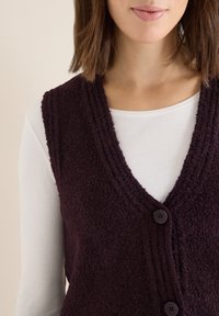 Burgundowy kamizelka o fakturze z dekoltem w kształcie litery V, dwoma guzikami i prążkowanymi detalami, nałożona na białą koszulę z długim rękawem.