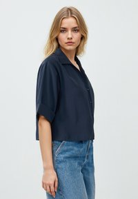 Camicia button-up a maniche corte blu navy con taglio morbido, colletto standard e tessuto liscio, abbinata a jeans a vita alta di un blu chiaro.