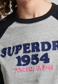 Grauer Kapuzenpullover mit marineblauen Raglanärmeln. Mit gesticktem Text "SUPERDRY 1954 TOKIO, JAPAN" in Blau und Pink.