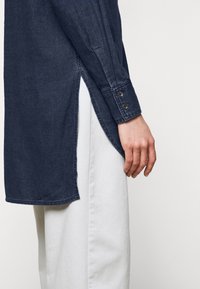 Vue de profil d'une personne portant une chemise à manches longues bleu foncé avec un ourlet fendu sur un pantalon clair.