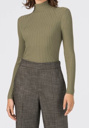 Nainen, joka pukeutuu oliivinvihreään, ribbineulottuun poolopaitaan ja korkeavyötäröisiin ruskeisiin ja mustiin haukkakuvioituihin housuihin.