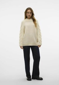 Cremefarbener Strickpullover mit hohem Kragen und geflochtenen Details an den Ärmeln, kombiniert mit dunkelblauen Jeans und schwarzen Schuhen.